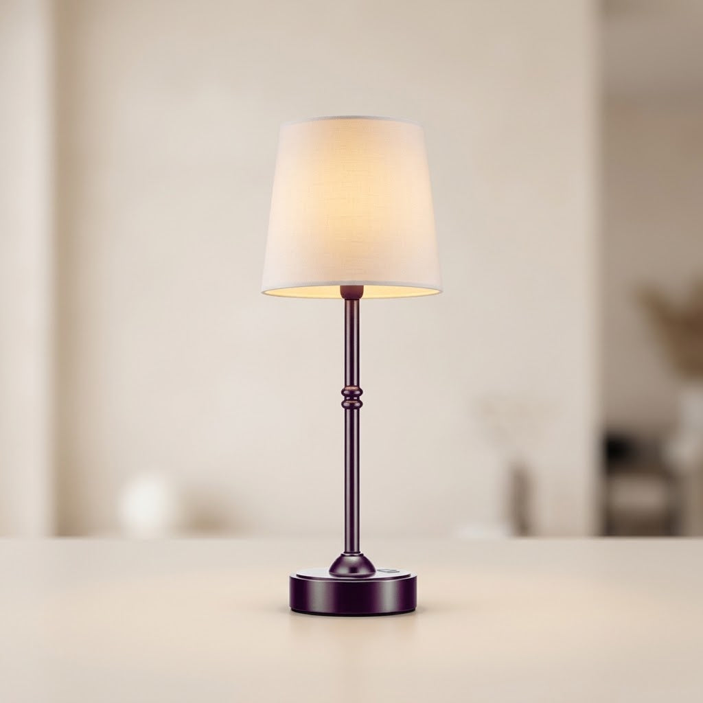 A small table lamp with a warm white fabric drum shade and a dark metallic base, shown lit on a light beige surface against a softly blurred indoor background.