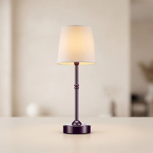 A small table lamp with a warm white fabric drum shade and a dark metallic base, shown lit on a light beige surface against a softly blurred indoor background.