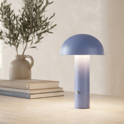 A modern mushroom-shaped table lamp in a dusty blue-gray color, glowing with a warm light on a light wood table in a softly blurred, minimalist living room setting.