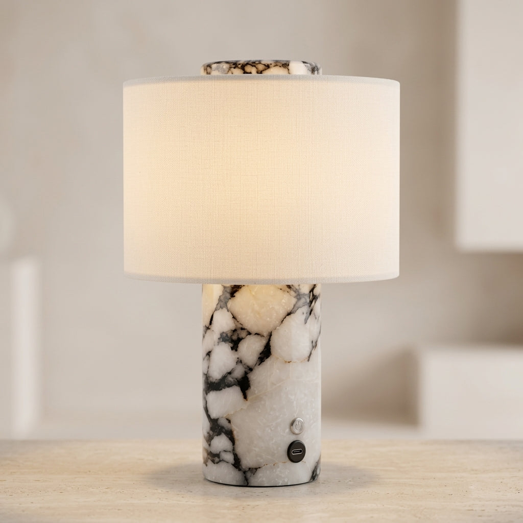 A modern table lamp with a white and black marbled stone base and a warm cream fabric drum shade, shown lit on a light-colored surface against a softly blurred neutral background.