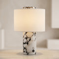 A modern table lamp with a white and black marbled stone base and a warm cream fabric drum shade, shown lit on a light-colored surface against a softly blurred neutral background.