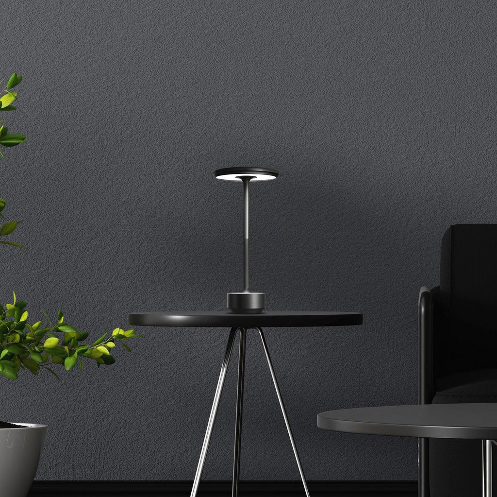 Modern black table lamp on a small round table with a dark gray wall and plant in the background.
