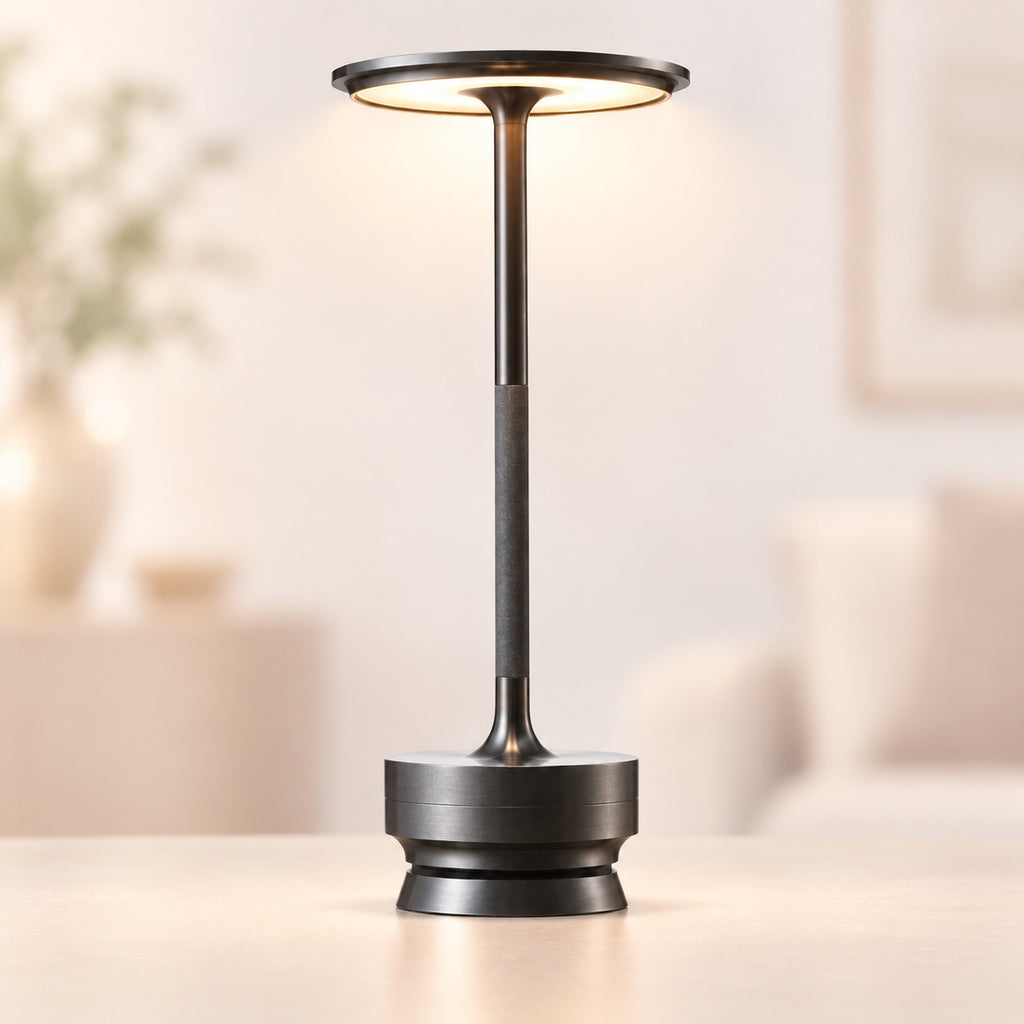 A modern adjustable table lamp with a dark metallic finish, featuring a circular head that emits warm light, standing on a round base on a light wooden table with a softly blurred living room background.