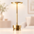 A modern gold table lamp with a circular shade, slim stem, and tiered base, shown lit on a light surface in a softly blurred indoor setting.